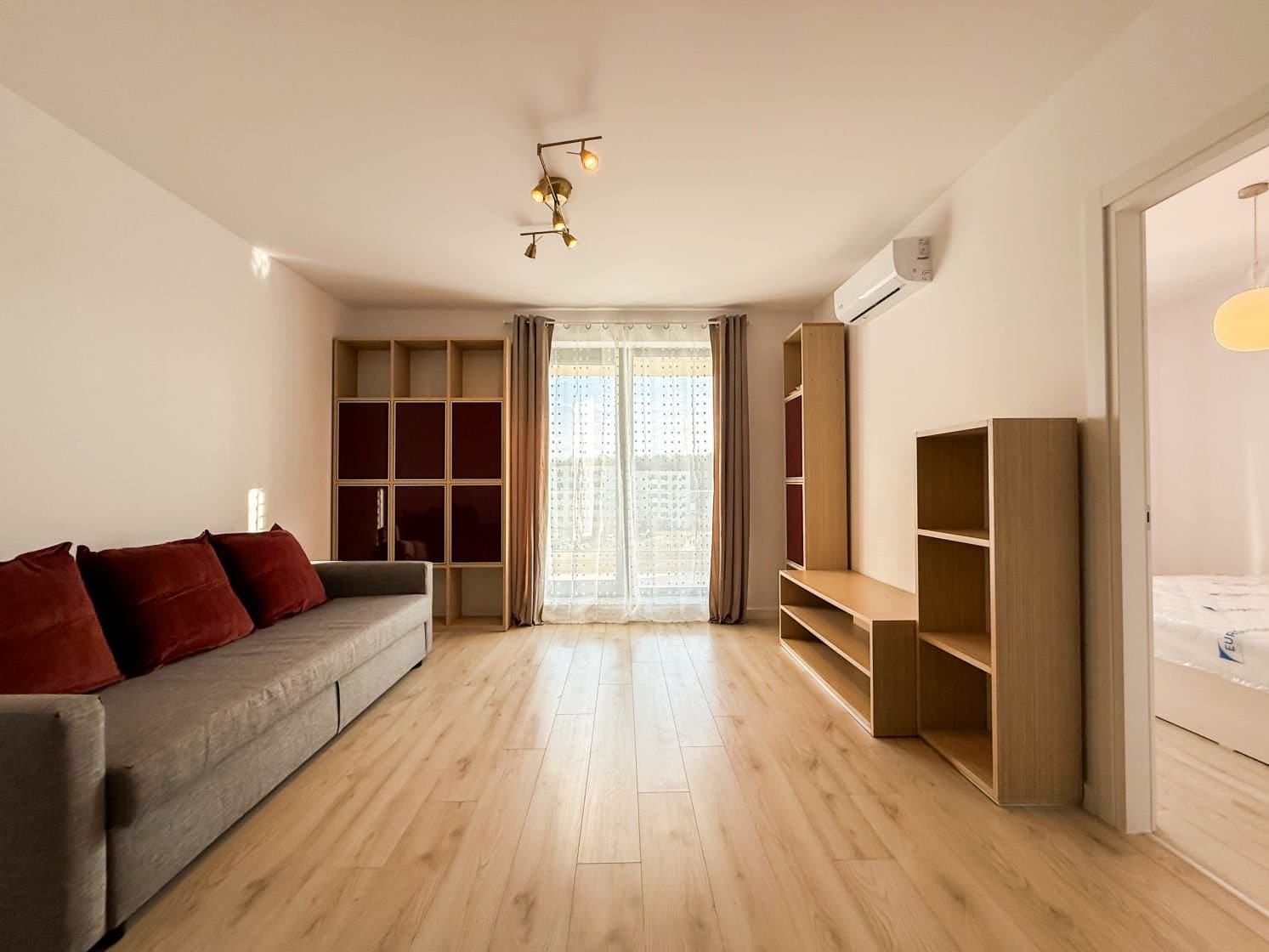 Living cu mobilă gri, perne roșii, parchet și balcon